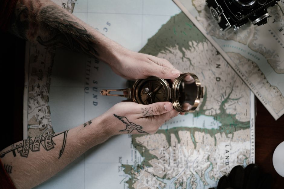 Hands holding a vintage compass over a map. Tattoos visible. Indoor shot.